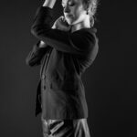 Schwarz-Weiß-Portrait von Alina im Studio: stehend im Blazer, die Arme über den Kopf gelegt