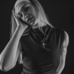Schwarz-Weiß-Portrait von Annika im Studio: sitzend im dunklen Kleid mit Haarband und Kreuzkette, Hand an der Wange