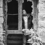 Schwarz-Weiß-Portrait von Paddy an einem Lost Place: Mann in Lederjacke lehnt im zerbrochenen Fenster eines alten Steinhauses